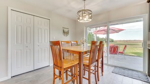 Uma área de refeições com mesa e cadeiras de madeira, um lustre e vista através de uma porta de vidro para um pátio externo com um guarda-sol vermelho.