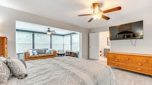 Quarto com uma cama grande, um ventilador de teto, cômoda de madeira, televisão na parede e um espaço de estar com sofá e poltronas.