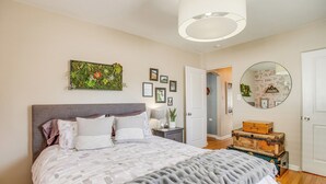Ein Schlafzimmer mit einem großen Bett, einem Nachttisch mit Lampe, einem Wandspiegel und einer Tür zu einem anderen Raum.