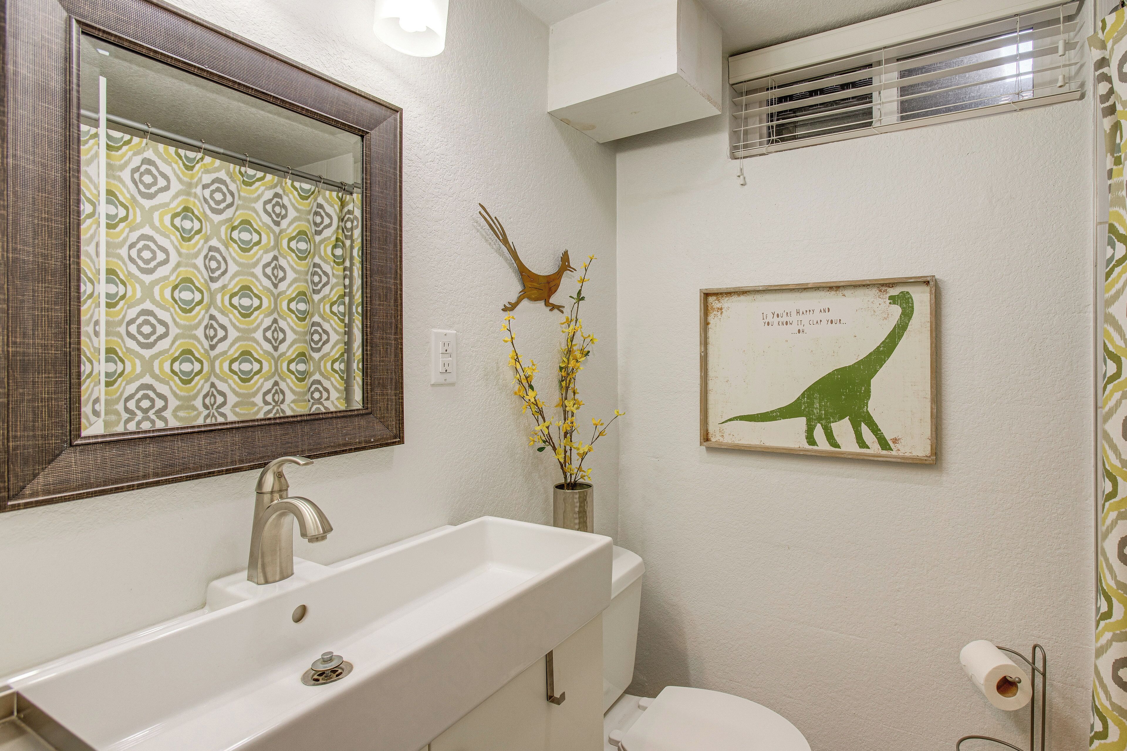 A bathroom with a white sink, mirror, and toilet. There is a framed picture of a dinosaur on the wall and a vase with yellow flowers.