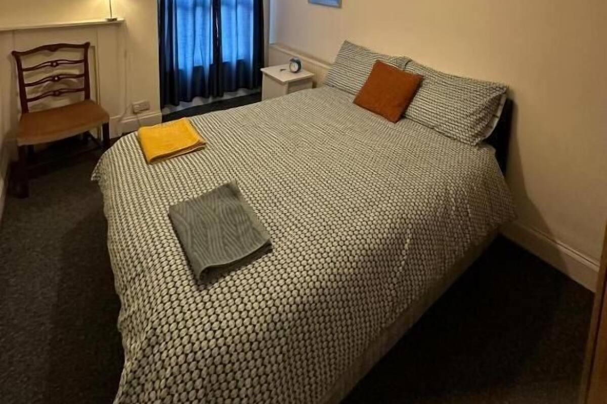 Quarto de hotel com cama, cadeira, mesa de cabeceira, abajur e janela com cortinas.