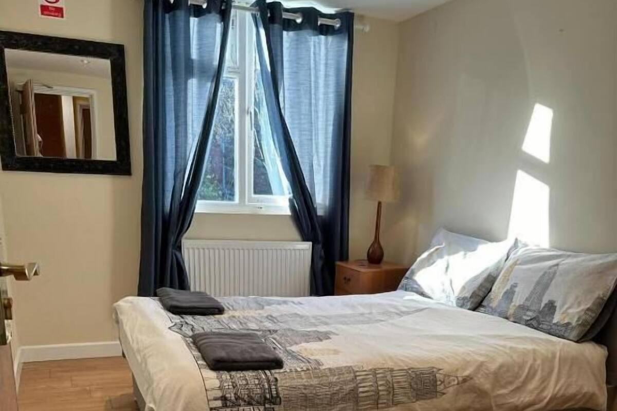 Quarto com cama, mesa de cabeceira, abajur, janela com cortinas e piso de madeira.