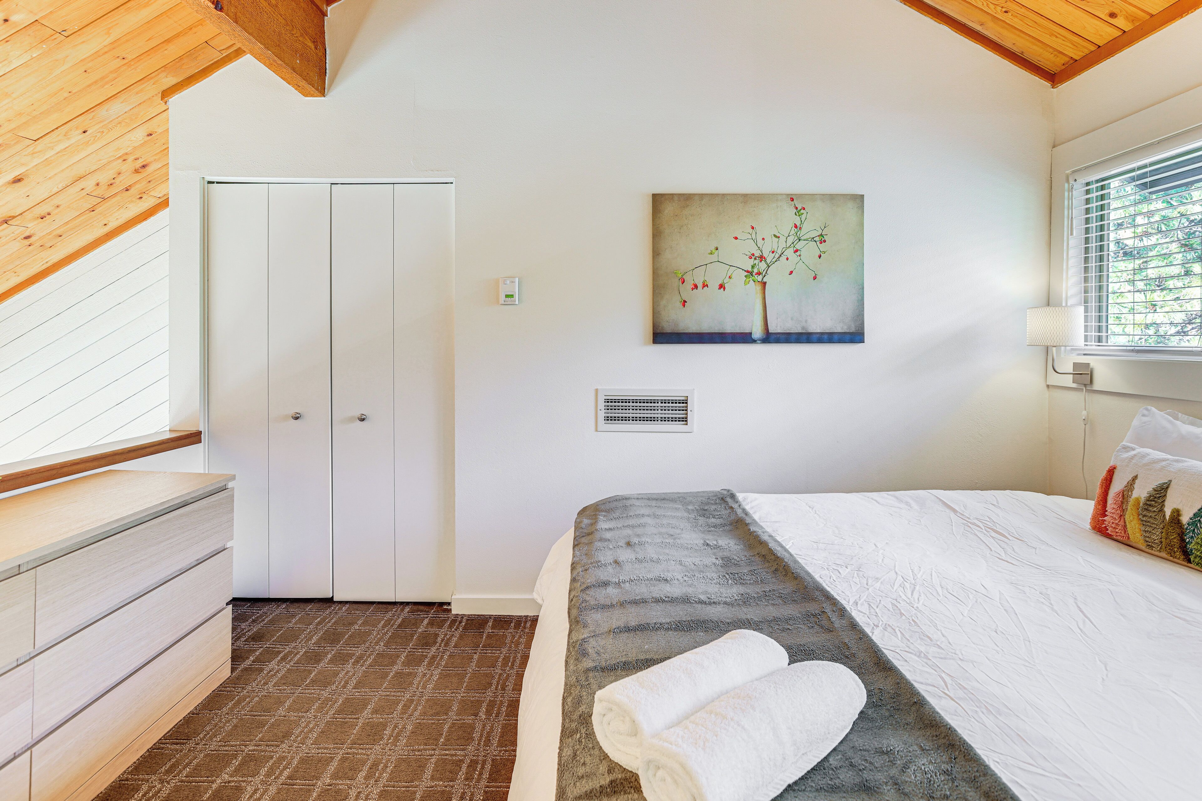 A bedroom with a bed, a painting, a window, and a wooden ceiling.