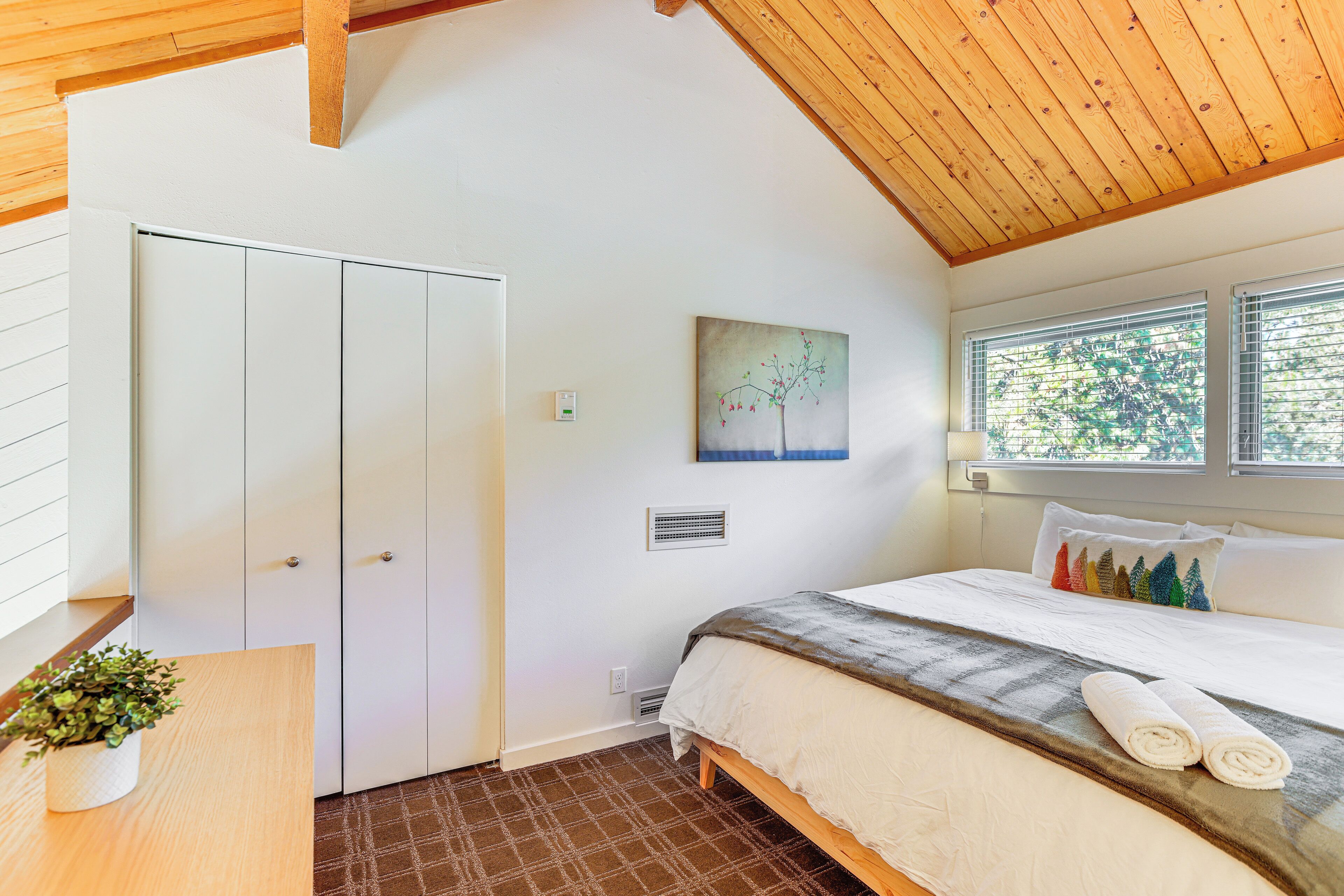 A bedroom with a bed, a bedside table with a plant, a painting on the wall, and a window with blinds.