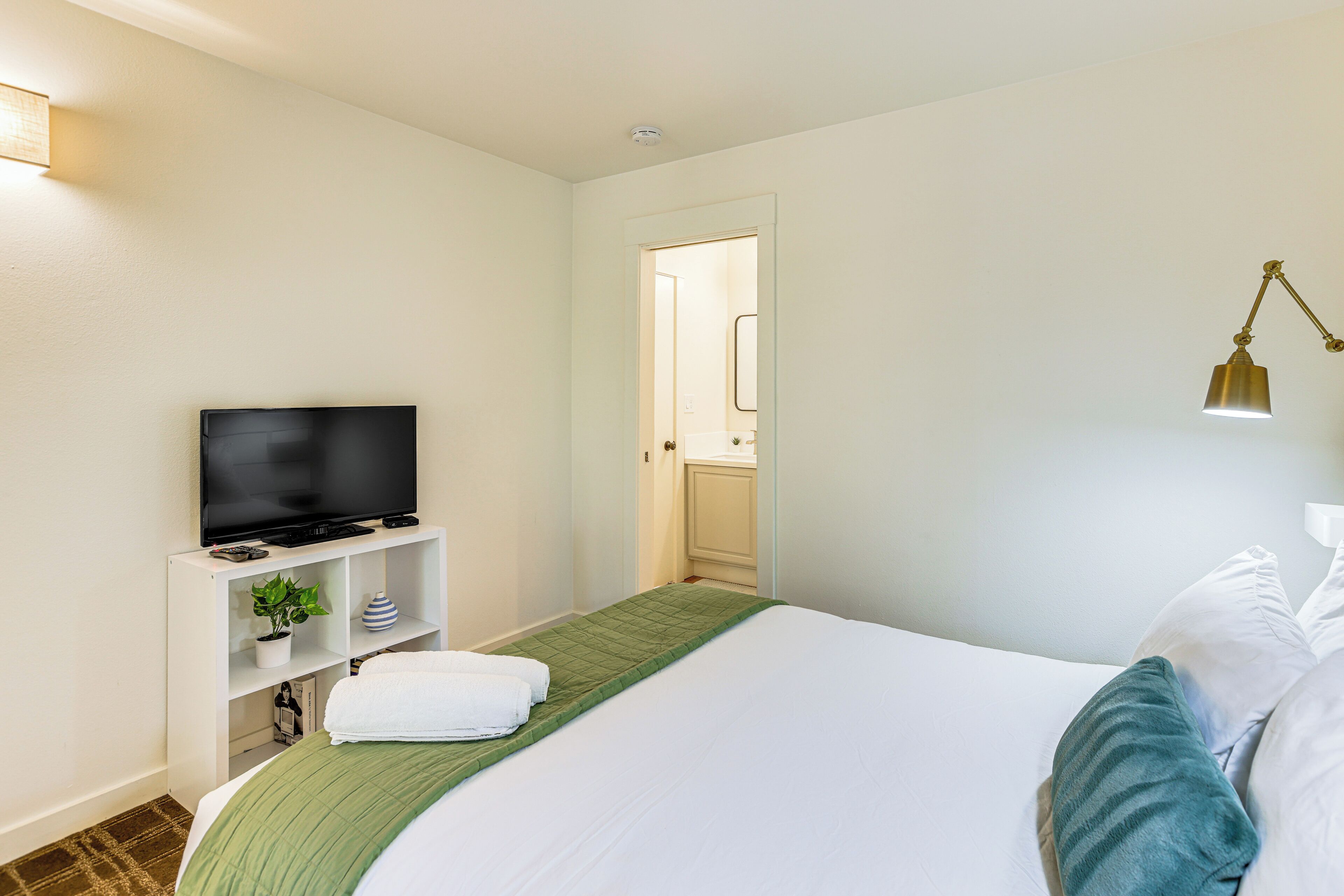 Een hotelkamer met een bed, een televisie op een standaard, een groene dekbedovertrek, witte kussens en een kleine plant op een plank.
