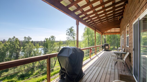 A covered deck with a grill, outdoor seating, and a view of a lake and forest. - Kenai River Views & Access: Soldotna Apartment! (Soldotna)