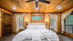 Een slaapkamer in een houten blokhut met een groot bed, twee nachtkastjes, een plafondventilator en een raam met gordijnen.