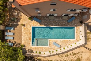 Vista aérea de una piscina con solárium de piedra y azulejos, rodeada de sillas de descanso y un pequeño jardín.
