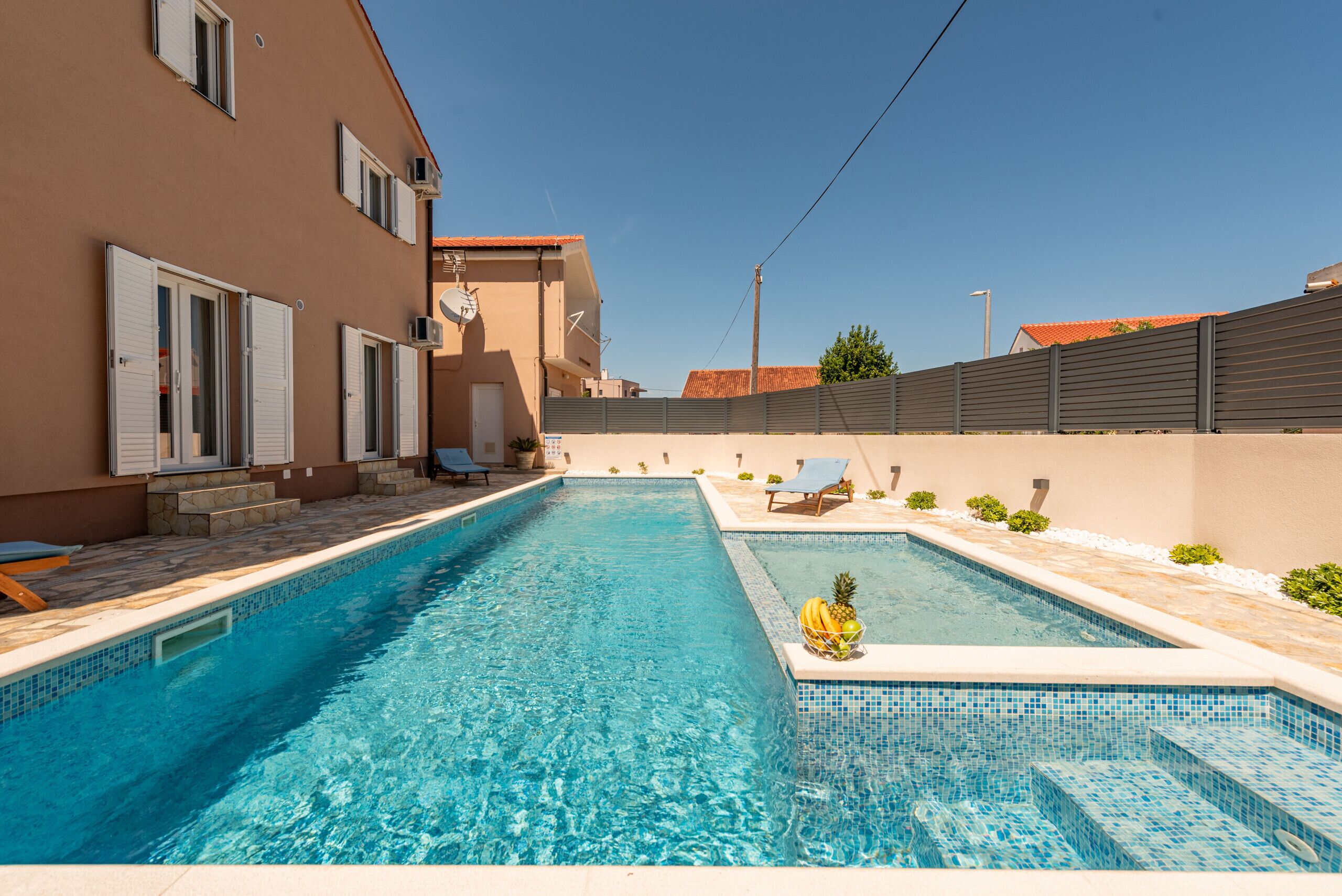 Ein Swimmingpool mit klarem, blauem Wasser, umgeben von einem beige gefärbten Gebäude mit weißen Fensterläden und einer gefliesten Terrasse.