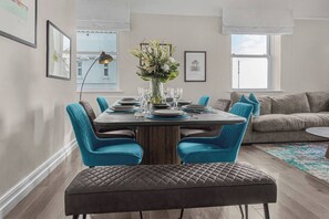 A dining area with a rectangular table set for four, surrounded by teal upholstered chairs. A vase with flowers is on the table, and there is a window with a view of buildings in the background.