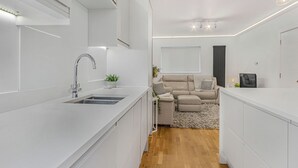 Ett modernt kök med vita skåp, en diskho i rostfritt stål och en inbyggd ugn. Det finns en matplats med ett vitt bord och stolar, och ett vardagsrum med en beige soffa och en grå matta.