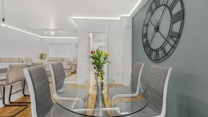 Ett modernt matområde med ett glasbord, grått klädda stolar och en stor väggklocka.