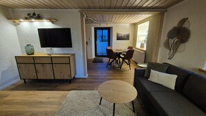 A modern living room with a sofa, a coffee table, a TV on a wooden cabinet, and a dining area with chairs and a table.