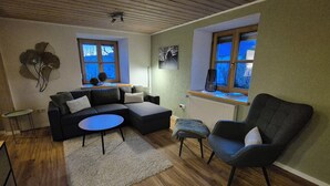 A cozy living room with a sectional sofa, a round coffee table, and a single armchair. There are two windows with blinds, a radiator, and a wall-mounted artwork.