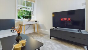 En moderne stue med et fladskærms-tv, der viser Netflix-logoet, et hvidt skrivebord med en lampe, en vase med blomster og et sofabord med to kopper.