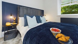 Et soveværelse med dybblåt sengetæppe, et natbord med lampe, en skål med jordbær og en tallerken med croissanter.
