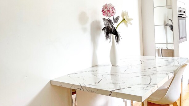 A white marble dining table with a white chair, a vase with flowers, and a kitchen in the background. - London 2-bed Apartment in Herbert Road (London)
