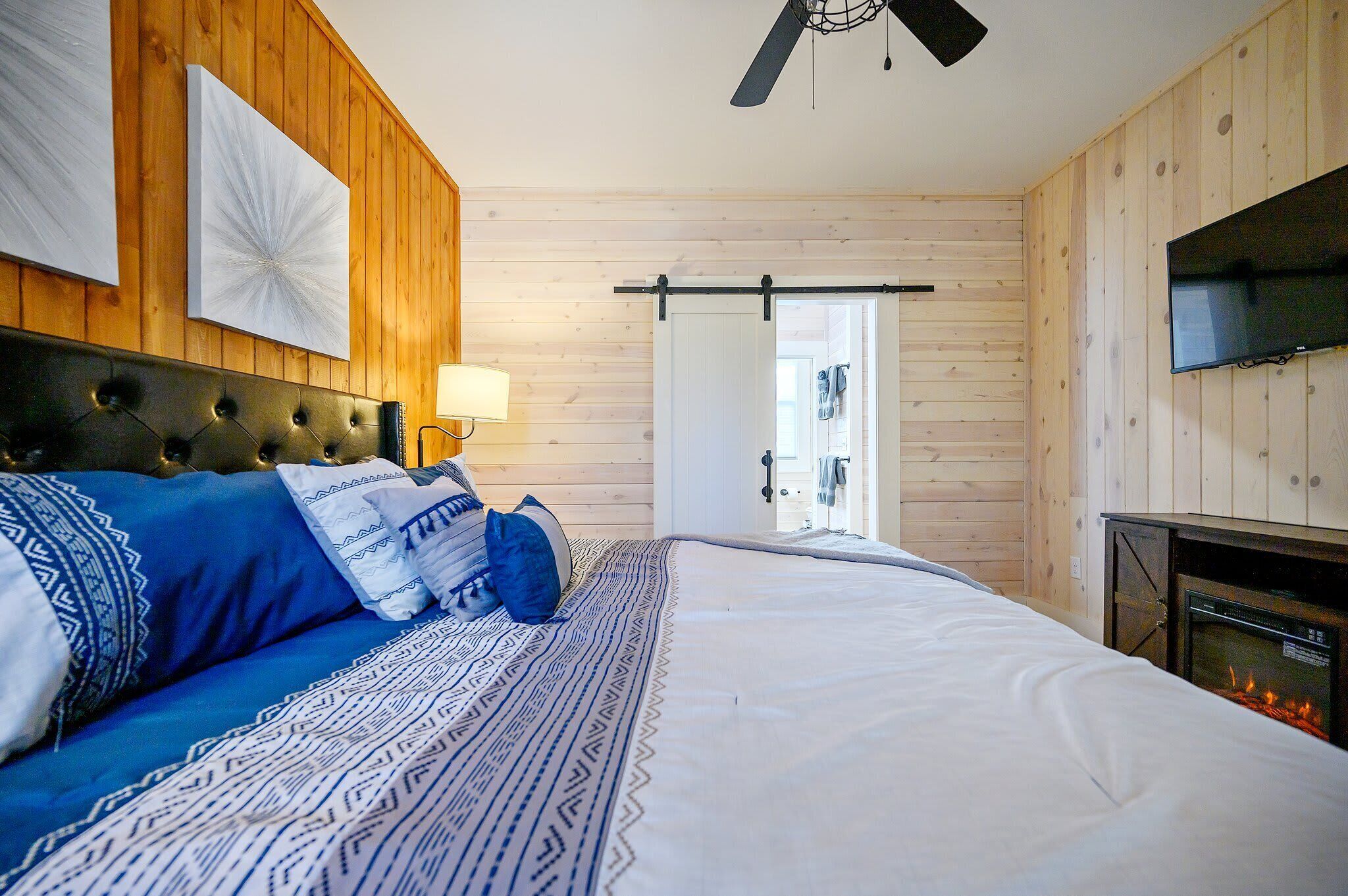 A bedroom with a large bed, a fireplace, wooden walls, a ceiling fan, and a TV.