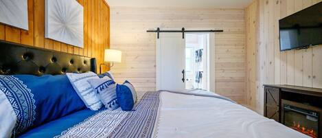 A bedroom with a large bed, a fireplace, wooden walls, a ceiling fan, and a TV.
