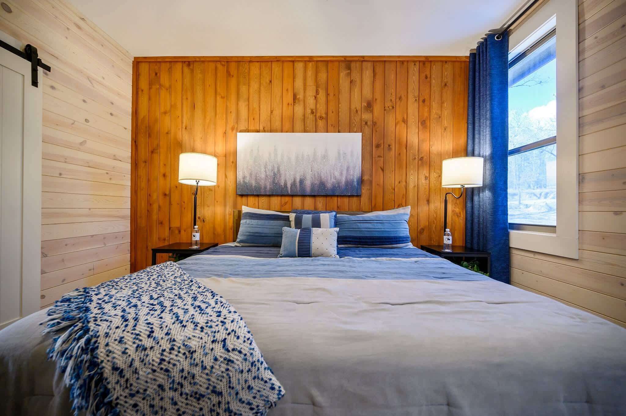 Ein Schlafzimmer mit einer Holzwand, einem großen Bett mit blauer Bettwäsche, zwei Nachttischlampen und einem Gemälde über dem Bett.