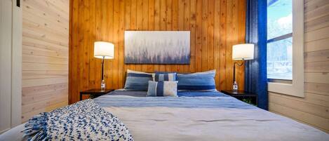 Ein Schlafzimmer mit einer Holzwand, einem großen Bett mit blauer Bettwäsche, zwei Nachttischlampen und einem Gemälde über dem Bett.