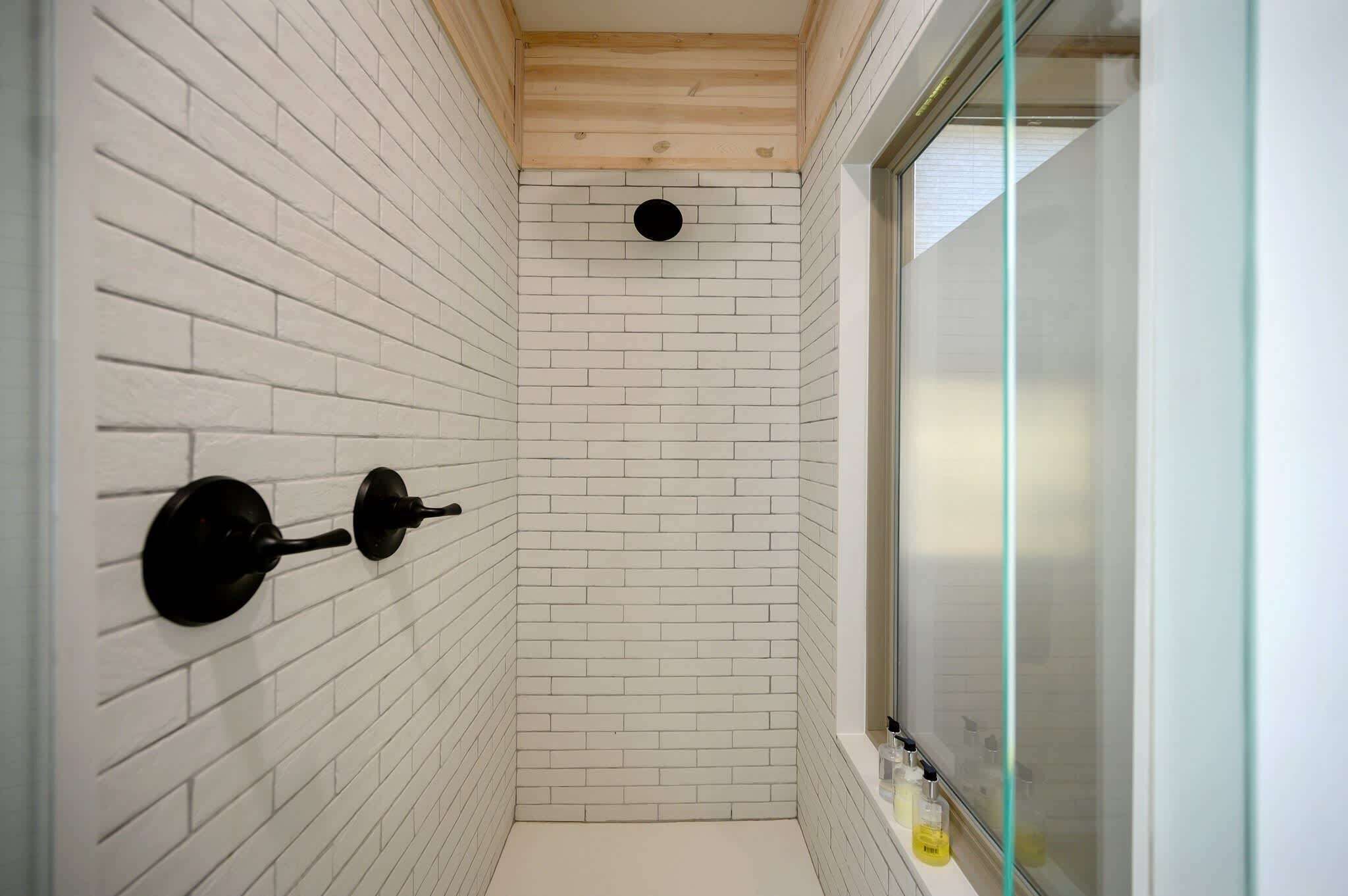 A narrow, white-brick-walled bathroom with a wooden ceiling and a glass shower enclosure.