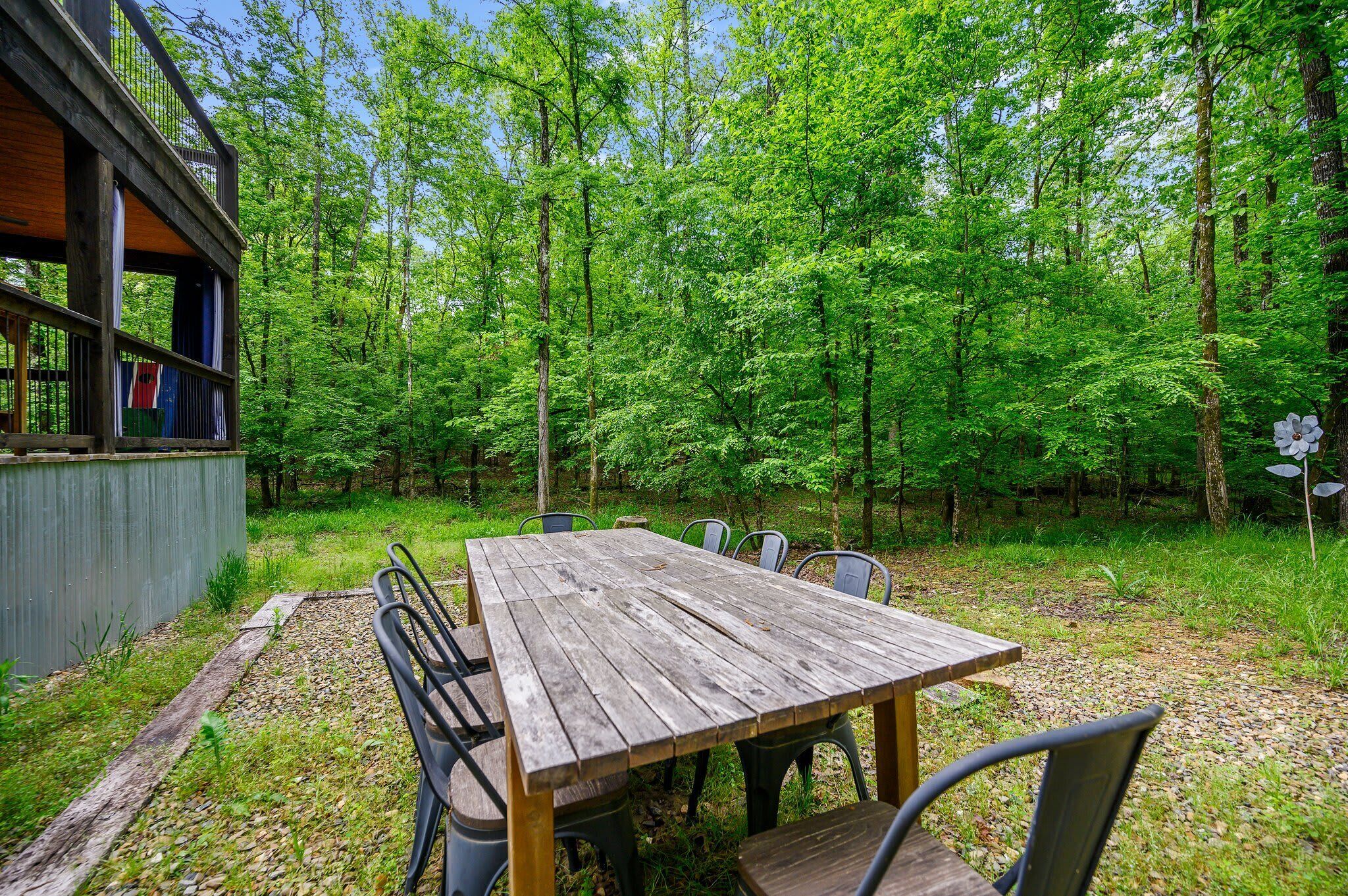 A rustic wooden table with metal chairs in a forested area, surrounded by trees and a wooden deck.