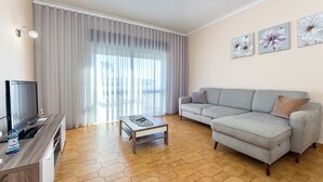 Una sala de estar con un sofá gris, una mesa de centro, un televisor y un ventanal con cortinas ligeras.