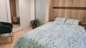 Quarto com uma cama grande, cabeceira de madeira, uma cadeira verde, uma mesinha e um vaso de planta.