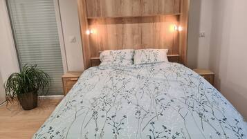 A bedroom with a wooden headboard, a floral bedspread, bedside lamps, and a potted plant.