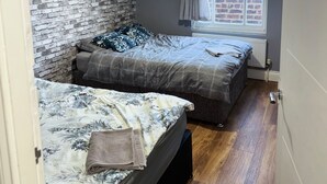 A bedroom with two beds, a brick wall, a window with curtains, and a hanging light fixture. - Westgate Haven - Spacious 4 Bedroom Apartment (Newcastle-upon-Tyne)