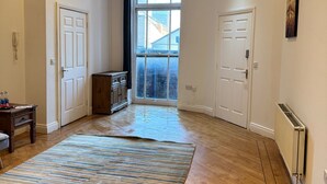 A spacious room with hardwood flooring, a large striped rug, and a view through a large window to the outside. - Westgate Haven - Spacious 4 Bedroom Apartment (Newcastle-upon-Tyne)