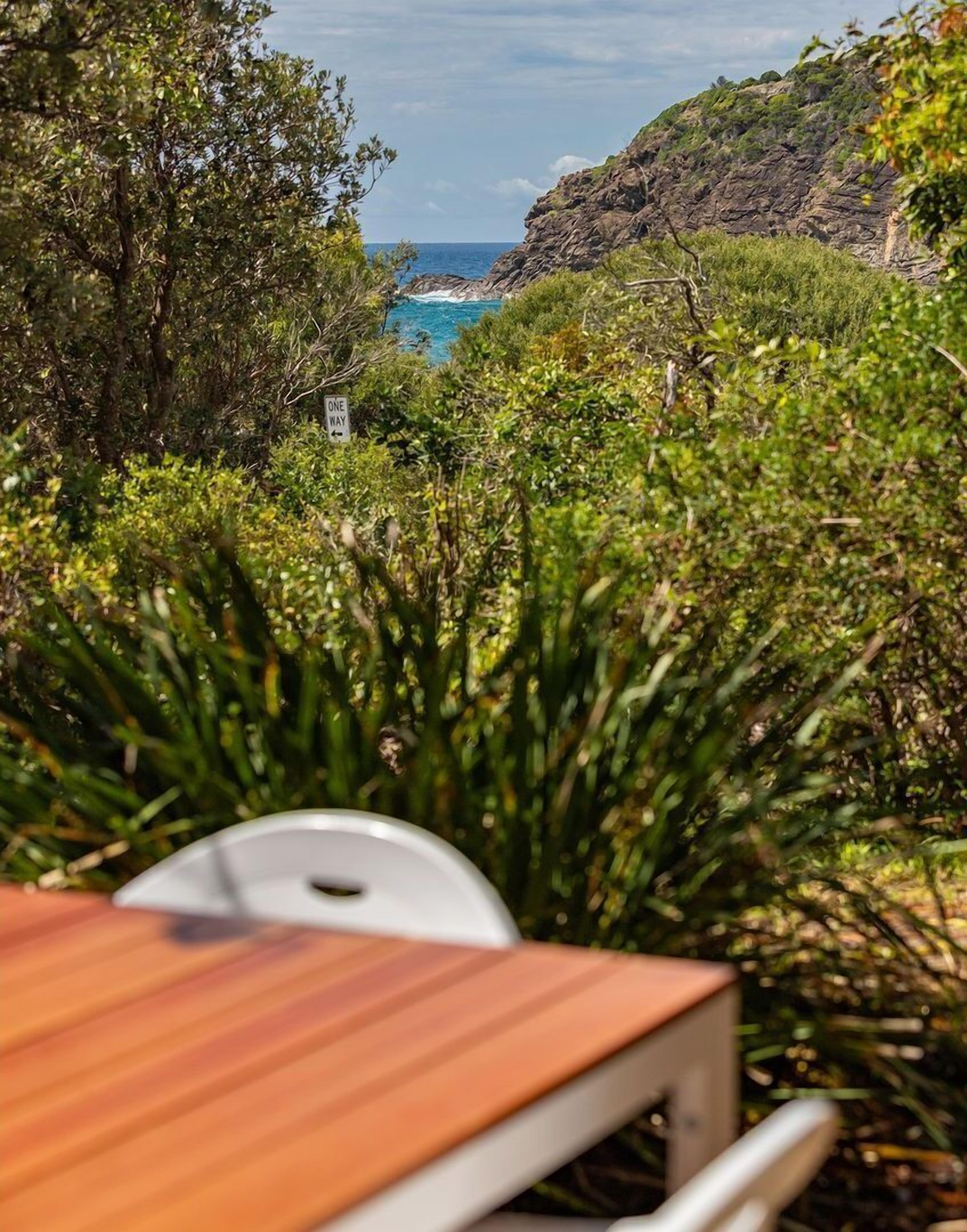A wooden table and white chair in a lush garden with a view of the ocean and rocky cliffs.