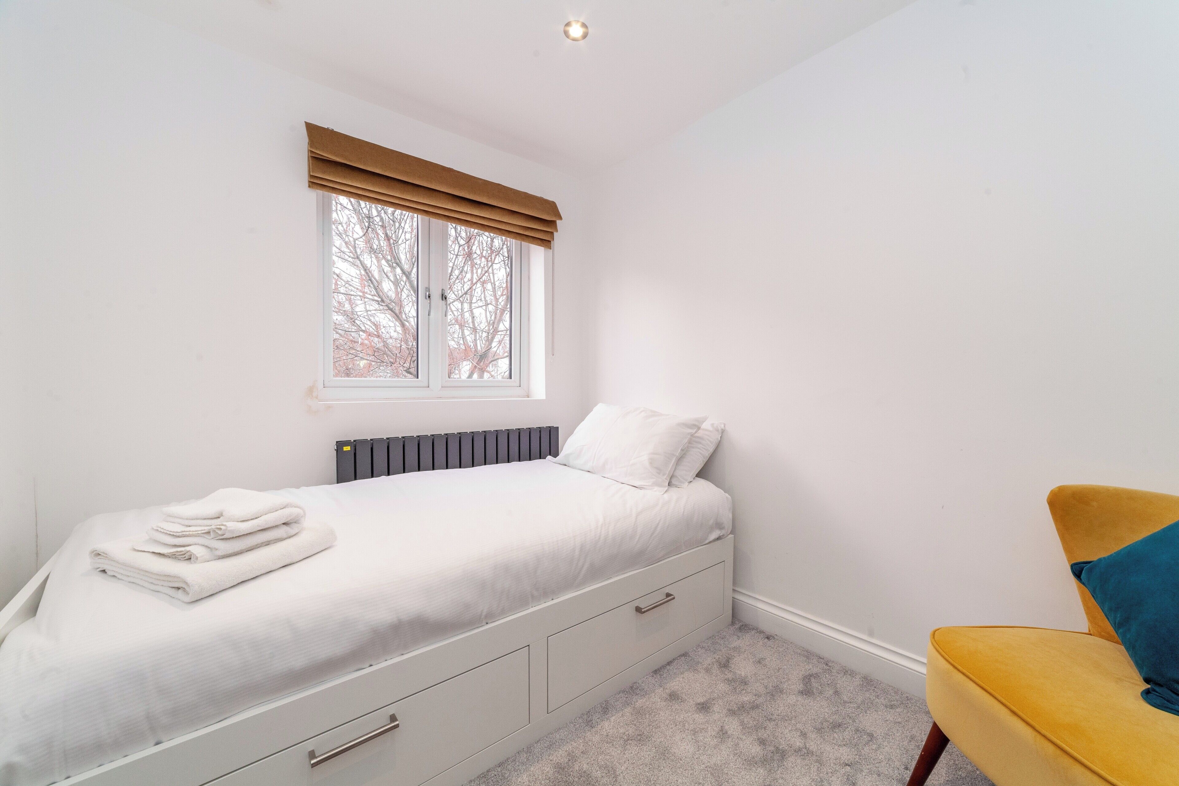 A bedroom with a bed, a window with blinds, a radiator, a chair, and a radiator on the wall.