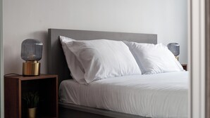 Un dormitorio moderno con cabecera gris, ropa de cama blanca y dos lámparas de noche sobre mesitas de madera.