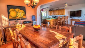 A spacious dining area with a wooden table and chairs, a fruit bowl, and a kitchen in the background.