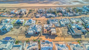 An aerial view of a coastal residential area with modern houses, some with flat roofs and others with domed structures, surrounded by sandy terrain and a beach in the background.