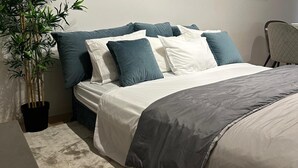 Una cama bien hecha con ropa de cama blanca y gris, una planta en maceta y una silla en una habitación de diseño sencillo y limpio.