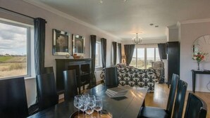 A dining area with a table set for six, a sofa in the background, and a view of the countryside through large windows. - Waterford Villa 5 Bed Beach Retreat (Ardmore)