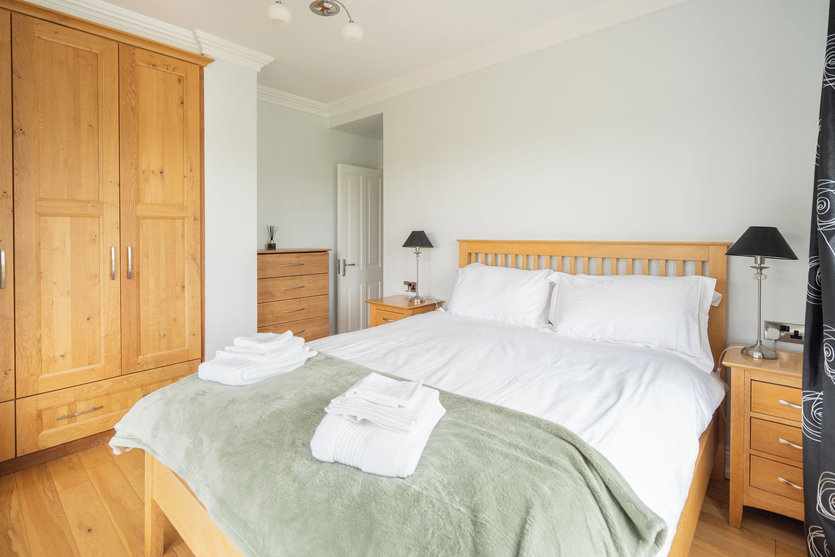 A bedroom with a large wooden wardrobe, a bed with white linens, two bedside tables with lamps, and a green throw blanket on the bed.