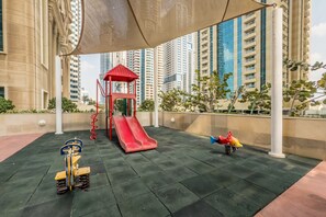 Une aire de jeux avec un toboggan rouge et une structure d’escalade, entourée d’immeubles de grande hauteur.
