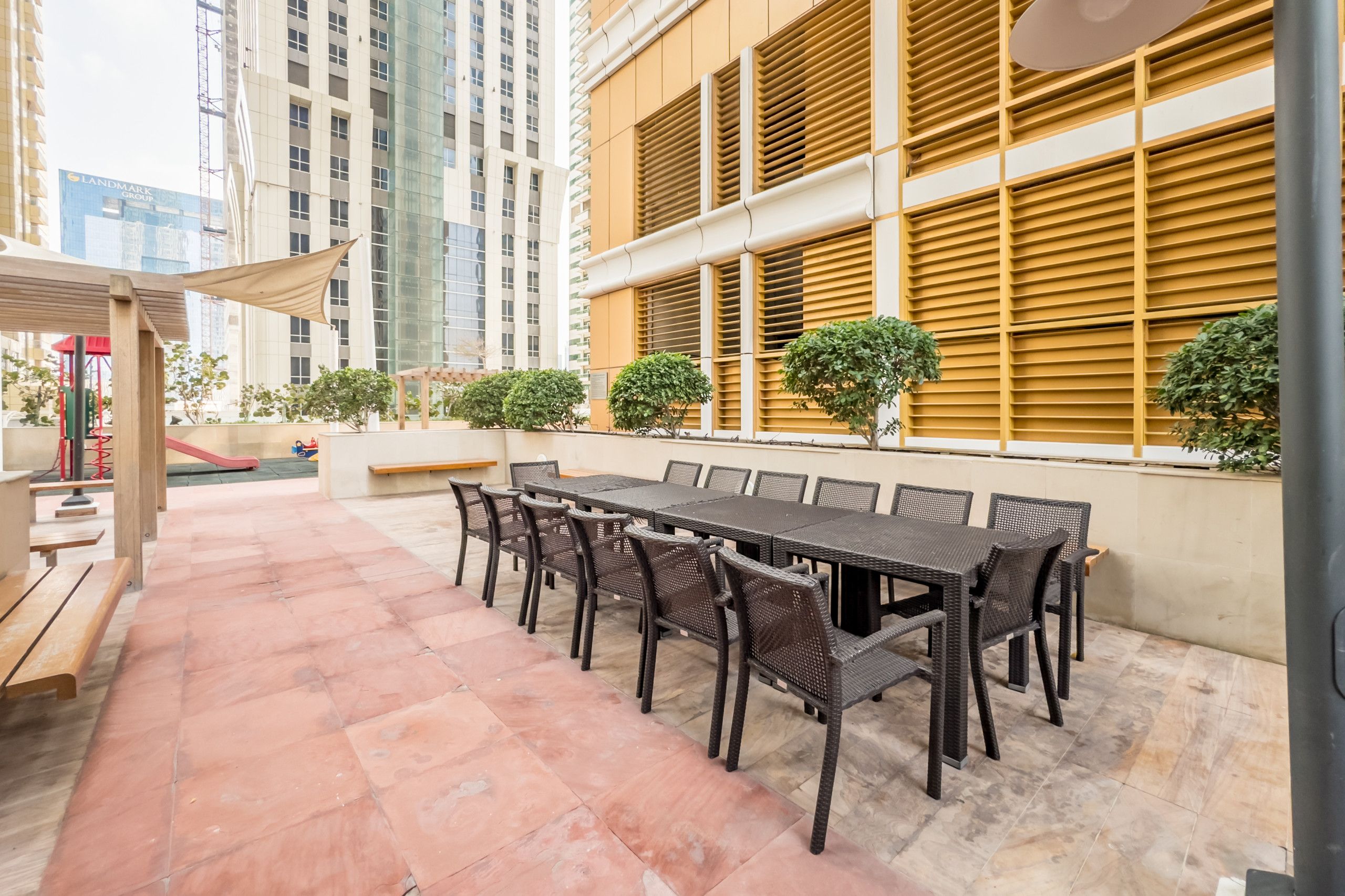 Une terrasse extérieure avec une longue table et des chaises, entourée d’un sol carrelé et d’un bâtiment doté de grandes fenêtres et de volets.