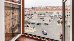 Vista desde una ventana sobre una concurrida calle urbana con edificios, una estatua y una parada de autobús.