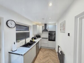 A compact kitchen with a sink, stove, and oven, a clock, and a window with blinds. - House Sleeps 5 With Garden and Parking (Birmingham)