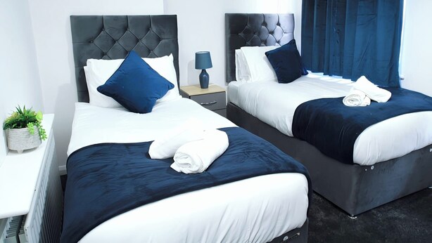 Ein Hotelzimmer mit zwei Betten, jeweils mit dunklem Kopfteil und weißer Bettwäsche. Ein kleiner Nachttisch mit Lampe und Topfpflanze ist ebenfalls vorhanden.