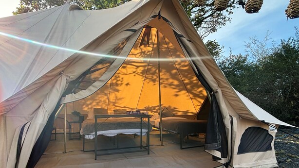 A tent with two beds and a table, set up on a rocky ground with trees in the background. - Cozy Hot Tent With Log Burner & Hot Tub Right Tent (Gaerwen)