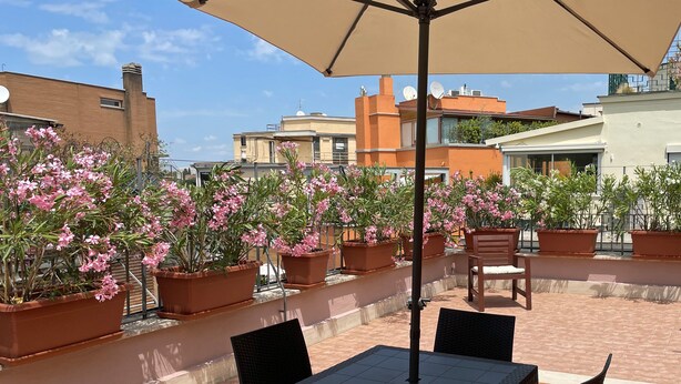 An outdoor patio with a table and chairs under a large umbrella, surrounded by potted plants with pink flowers. - Little Aventino B&B - Charming bed & breakfast with AC/WiFi in the heart of Rome (Rome)