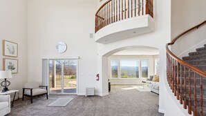 A spacious living room with a high ceiling, a staircase with wooden balusters, and a large window offering a view of the outdoors. - Stunning Mountain Retreat With Unmatched Views 5 Bedroom Estate (Oak Run)