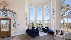 A spacious living room with large arched windows, a chandelier, and a wooden door with a glass panel. - Stunning Mountain Retreat With Unmatched Views 5 Bedroom Estate (Oak Run)
