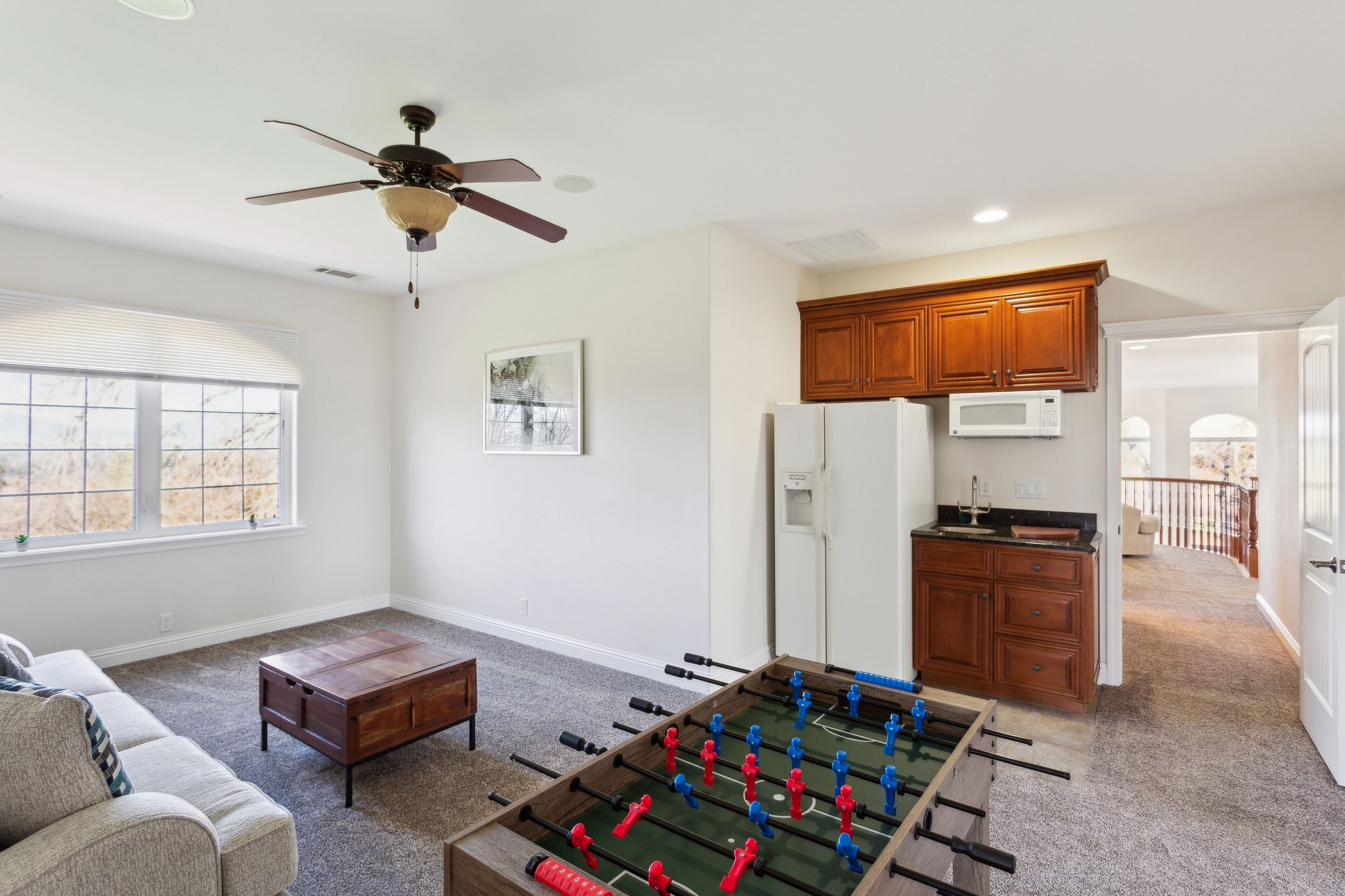 A living room with a foosball table, a ceiling fan, and a kitchen area with wooden cabinets and a microwave.